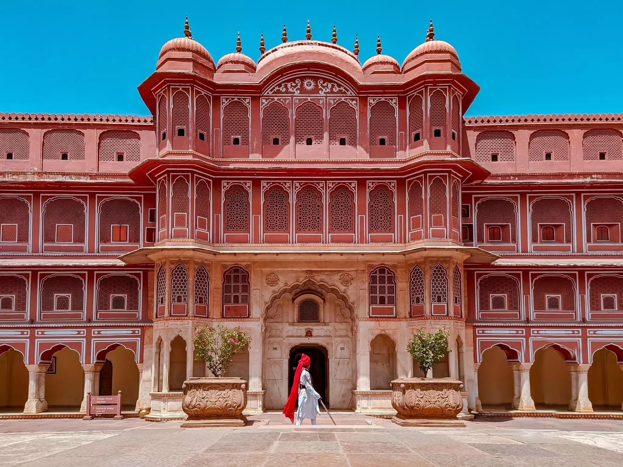 A person in white with a red head covering walks in front of a palace in India painted red with white detail around ornate windows.