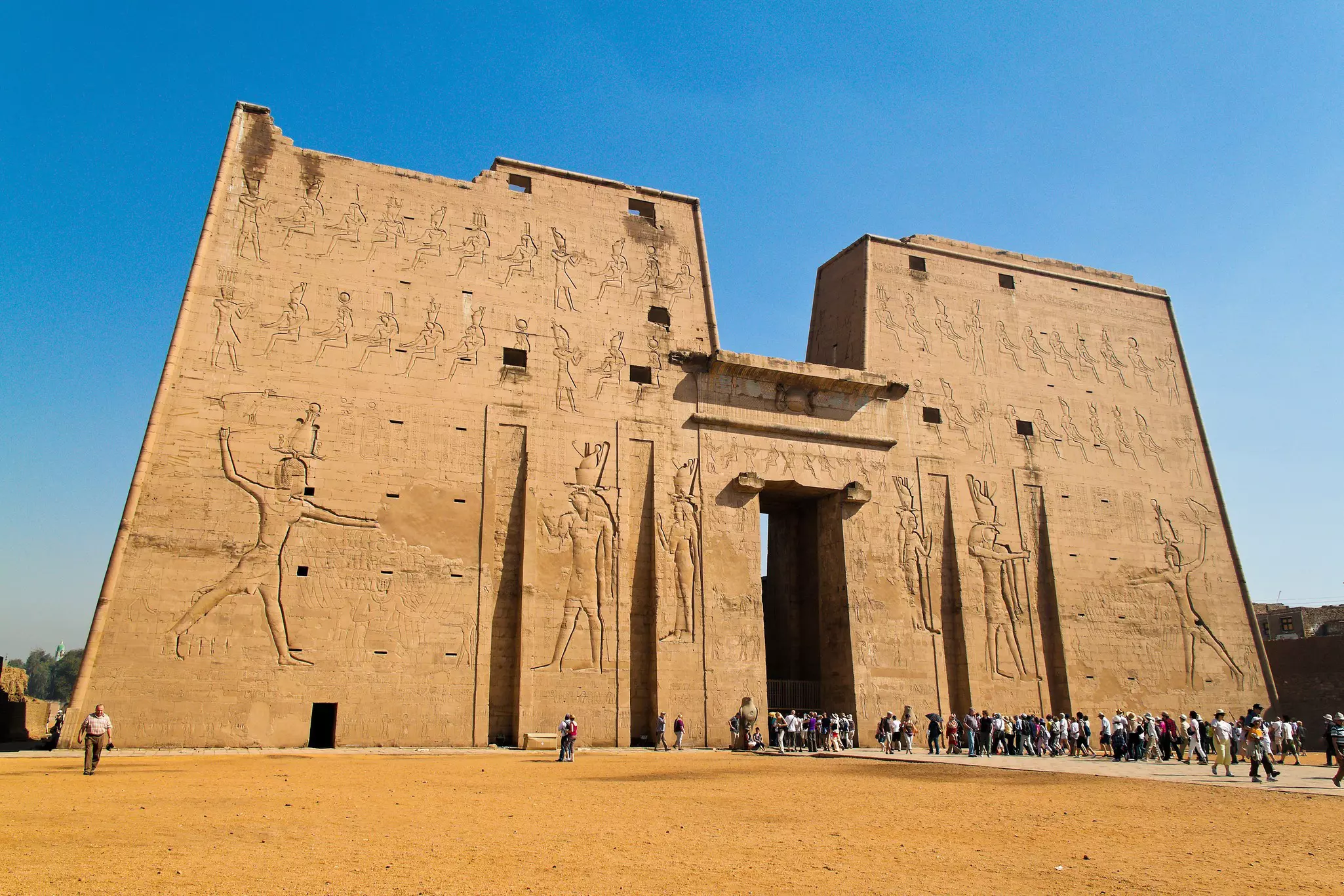 People enter a massive temple building relief sculptures of pharaohs on the exterior.
