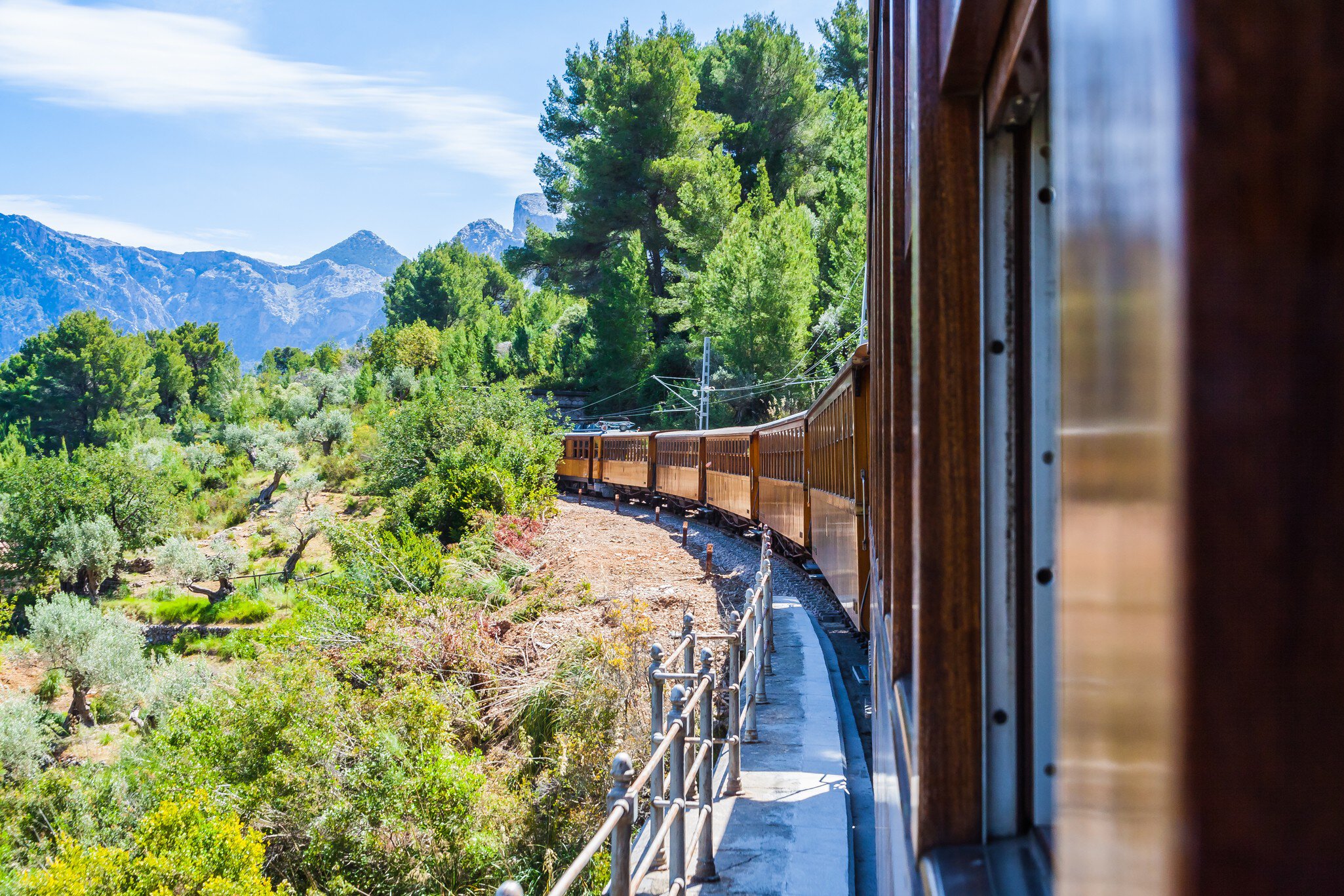 A vintage train with wooden carriages trundles through countryside and mountains on a sunny day.