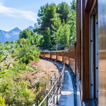 A vintage train with wooden carriages trundles through countryside and mountains on a sunny day.