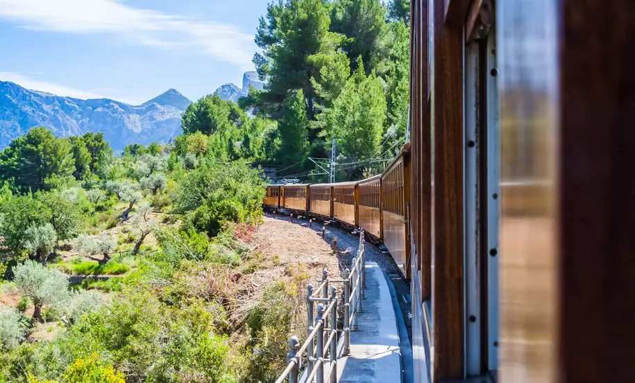 A vintage train with wooden carriages trundles through countryside and mountains on a sunny day.