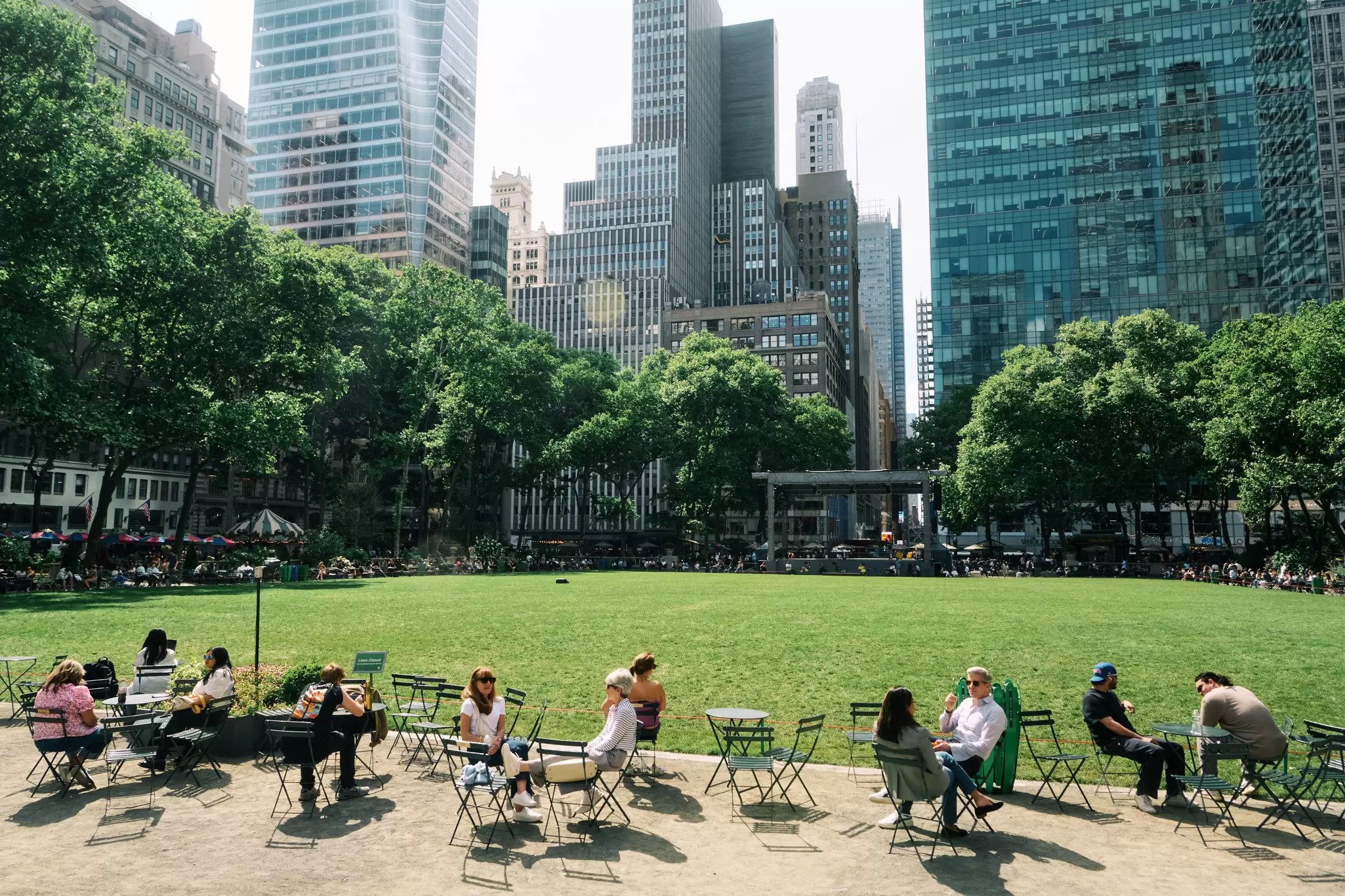 People sitting outside at tables near a big lawn and view of a city behind them.