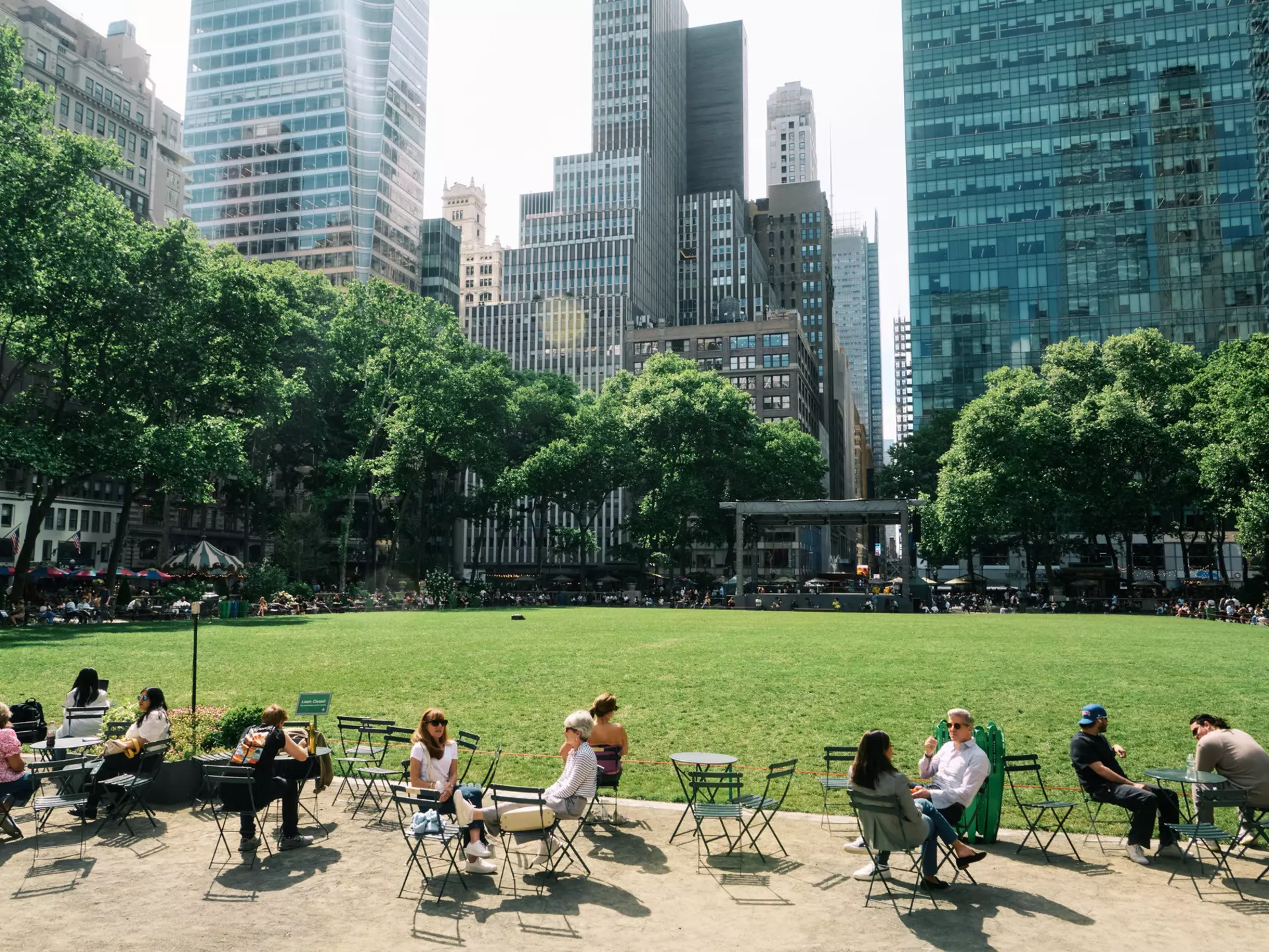 People sitting outside at tables near a big lawn and view of a city behind them.