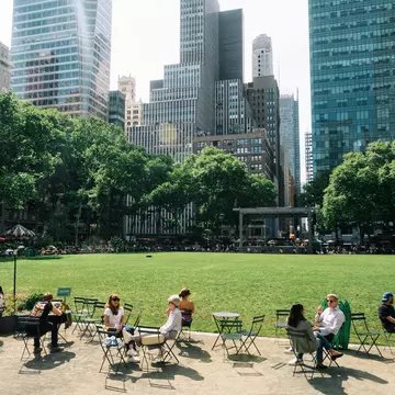 People sitting outside at tables near a big lawn and view of a city behind them.