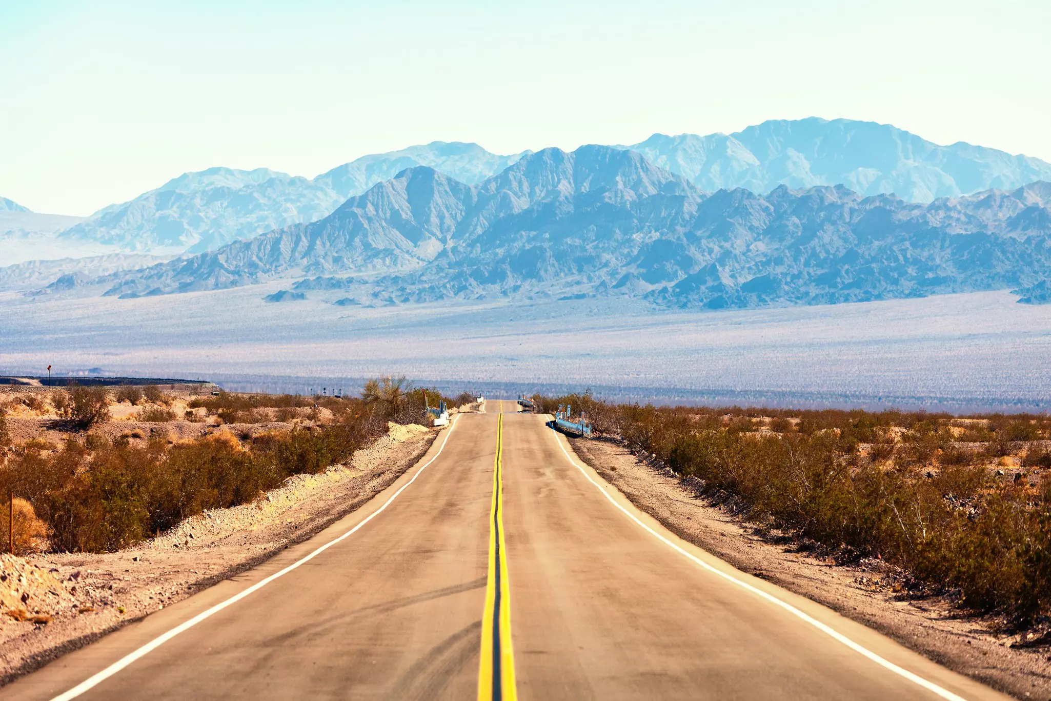 An empty road is seen passing through the desert in a foreshortened perspective. Mountains rise in the distance.