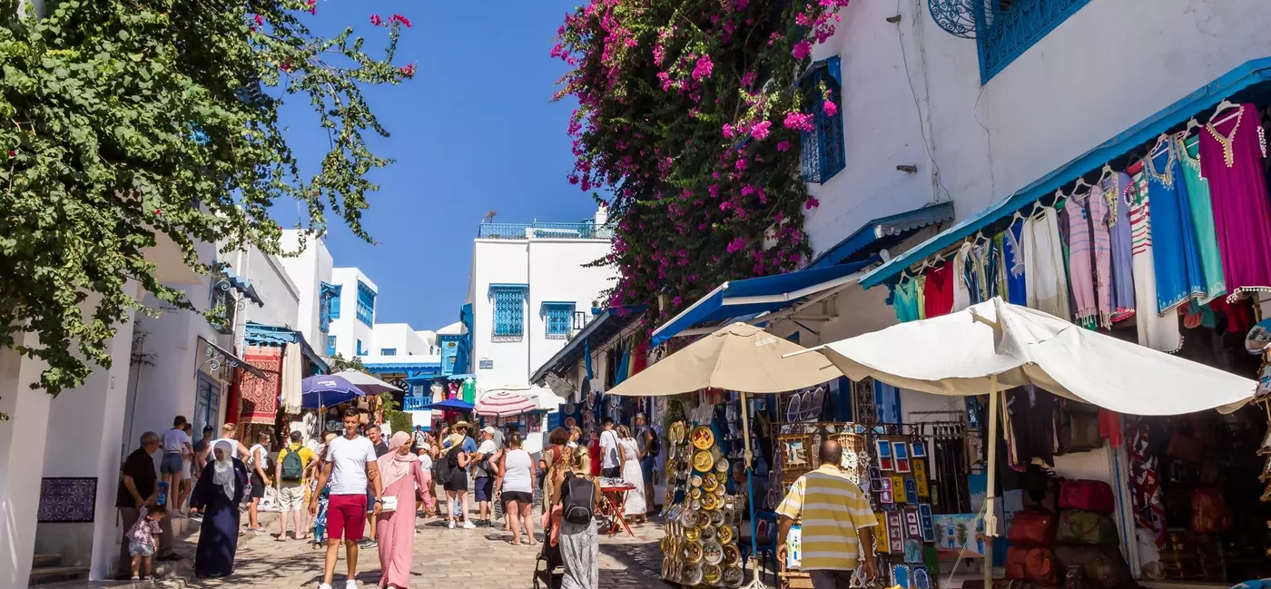 People walk down a street where outdoor stalls sell brightly colored clothes.