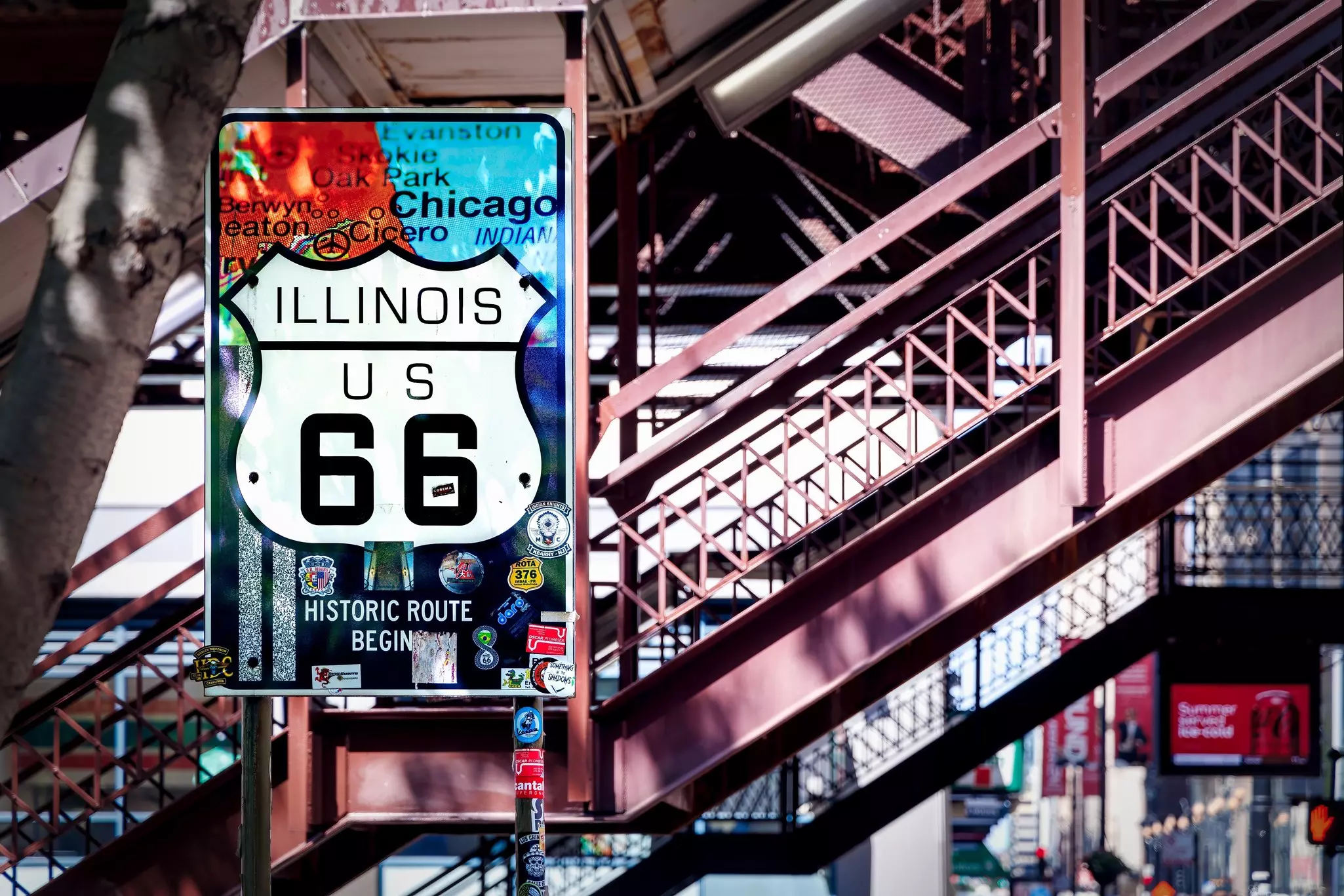A highway sign is displayed against a staircase leading to an elevated train in a city.