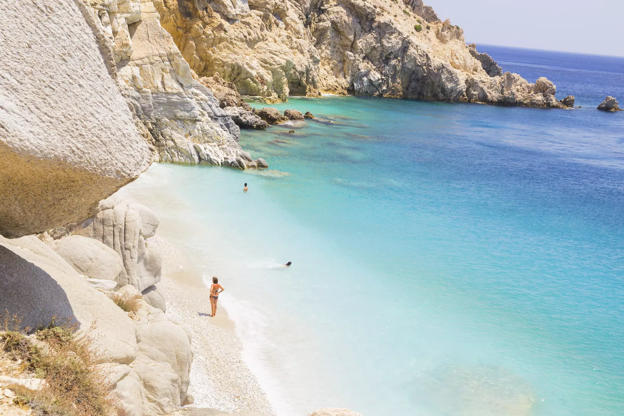 Beachgoers swim in clear waters while others walk on a white pebbly beach.