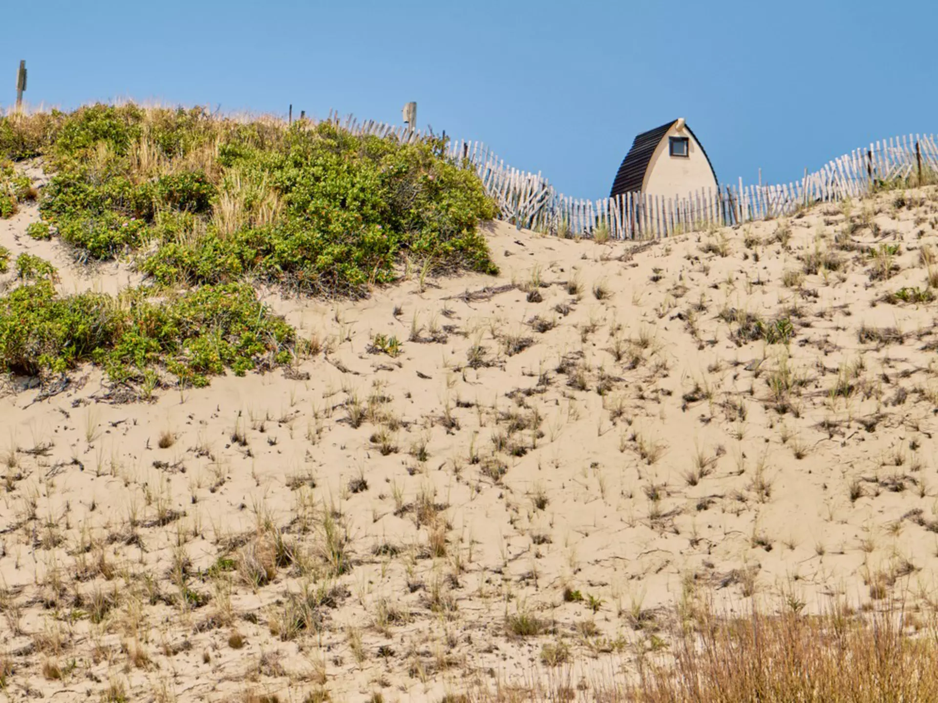 A structure at the top of a grassy and sandy dune under blue sky.