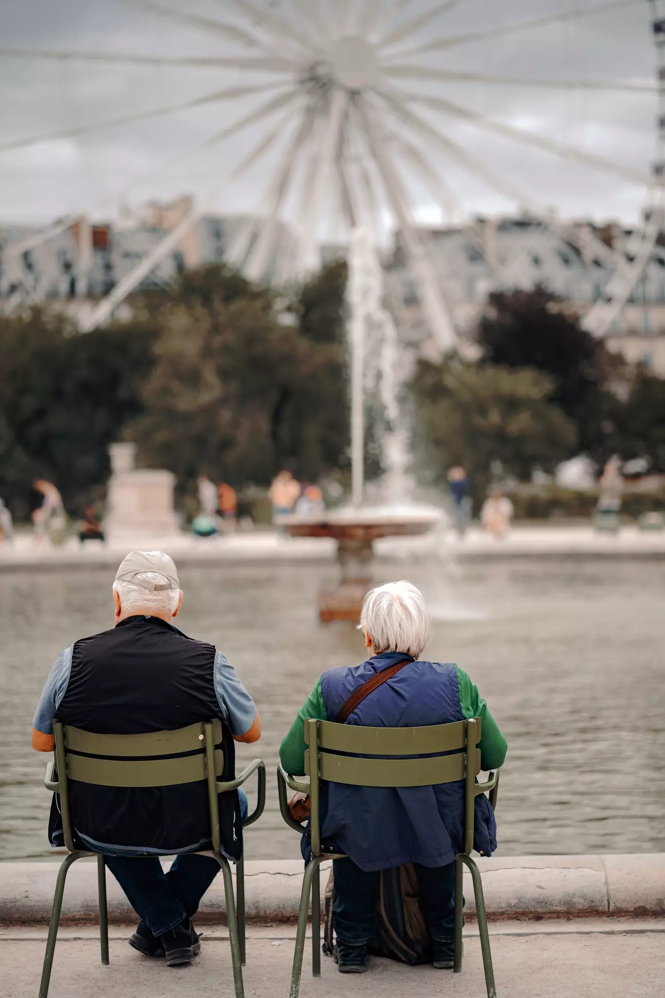 Paris, France - 13.07.2023: an elderly couple sits on green chairs next to a public spring fountain in a park in Paris,