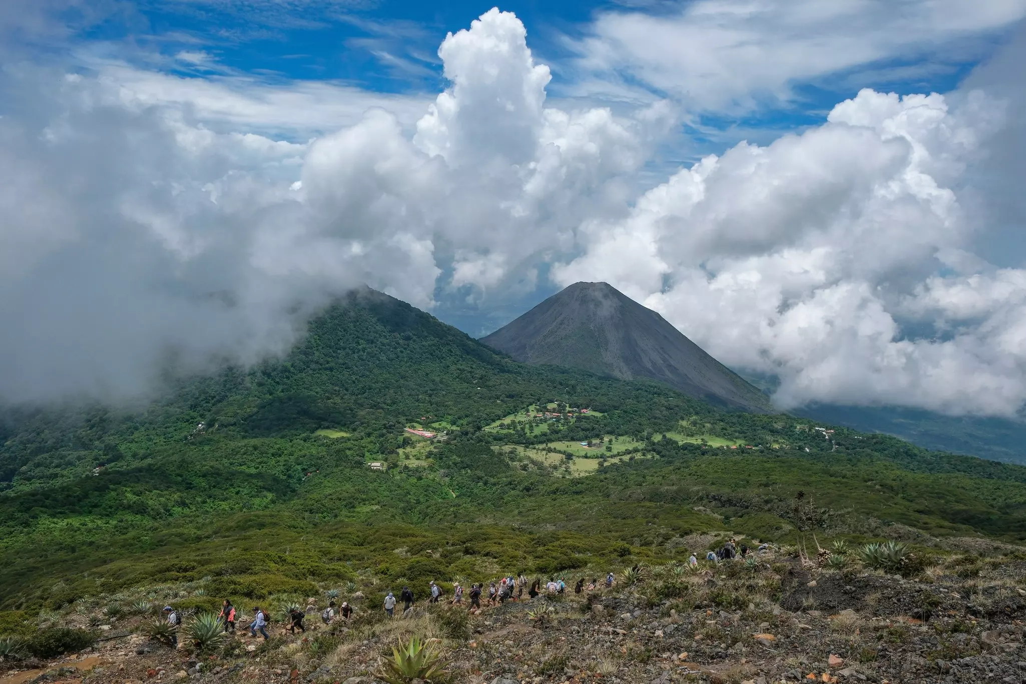 Hikers climb the rim of a volcano with other volcanoes in the background.