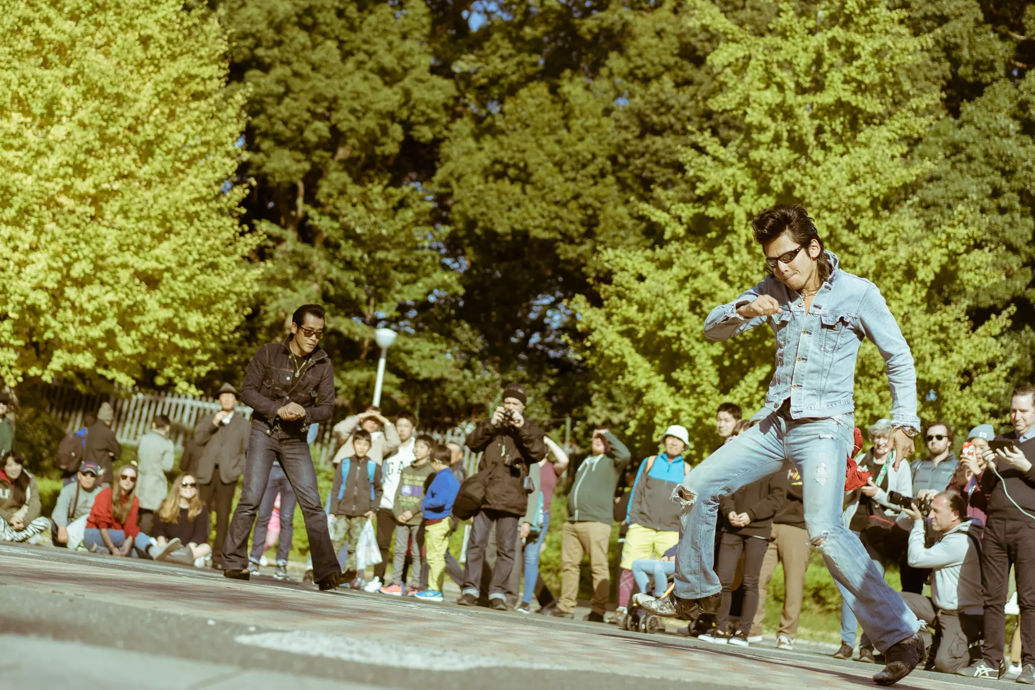 Men dressed as rock ’n’ roll performers dance in a city park as crowds watch.