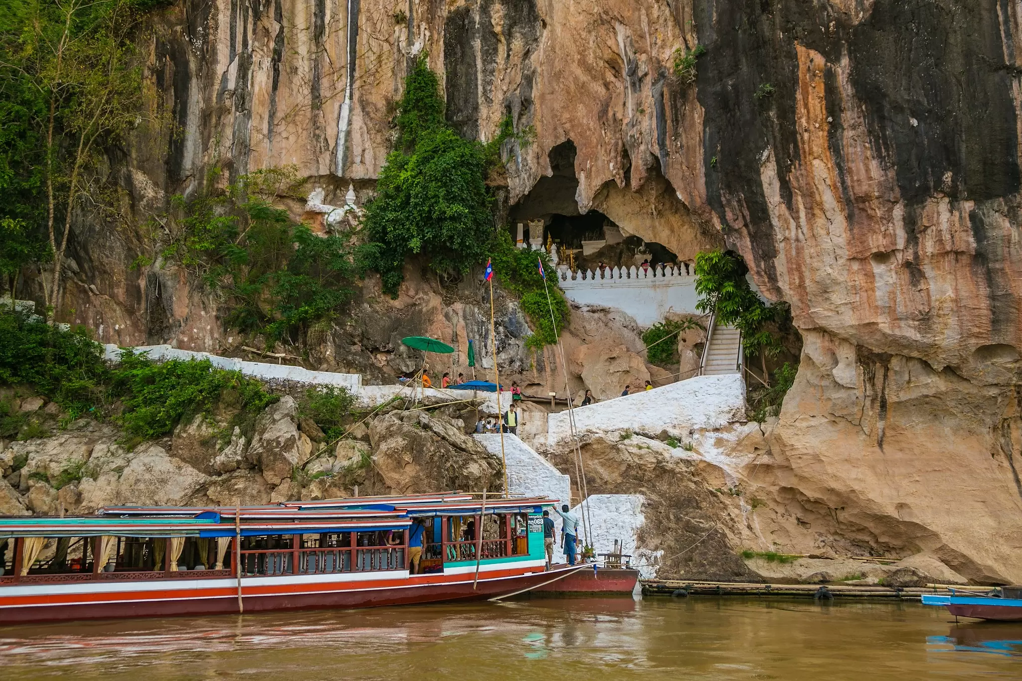Long wooden river boats moored below steep limestone cliffs and white steps leading up to a cave entrance.
