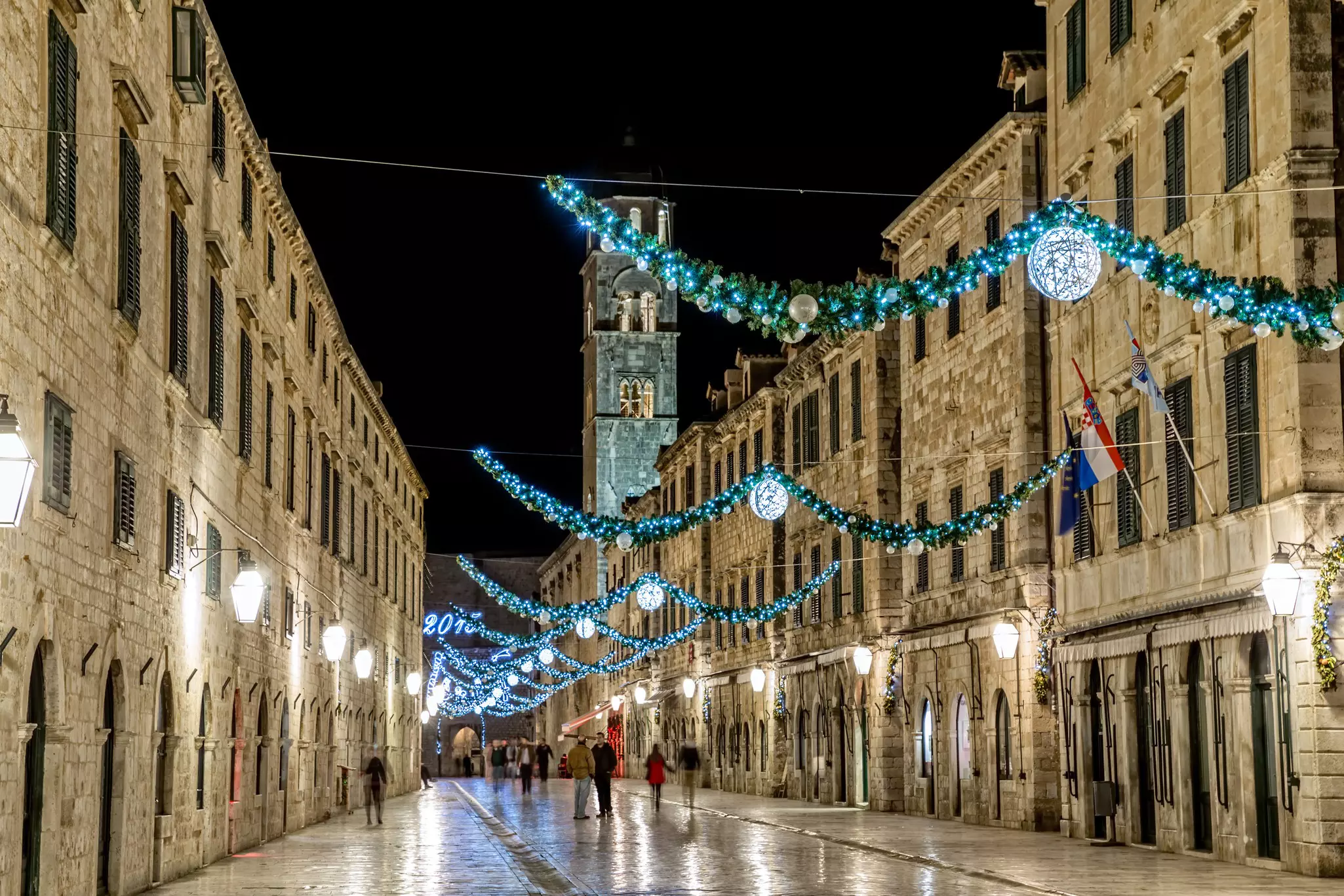 Festive holiday decorations hang above a pedestrian street in a historic city, pictured at night.