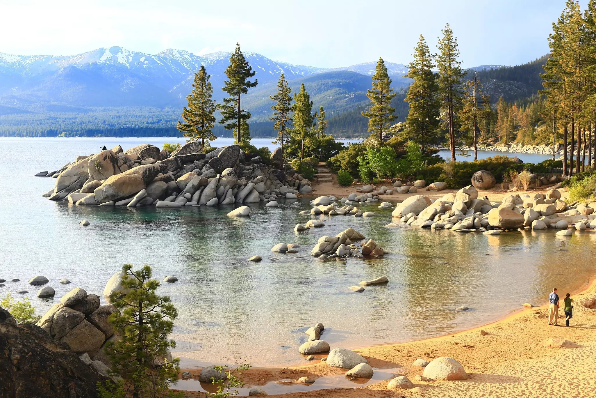 Two people walk on a sandy beach at the edge of an alpine lake.