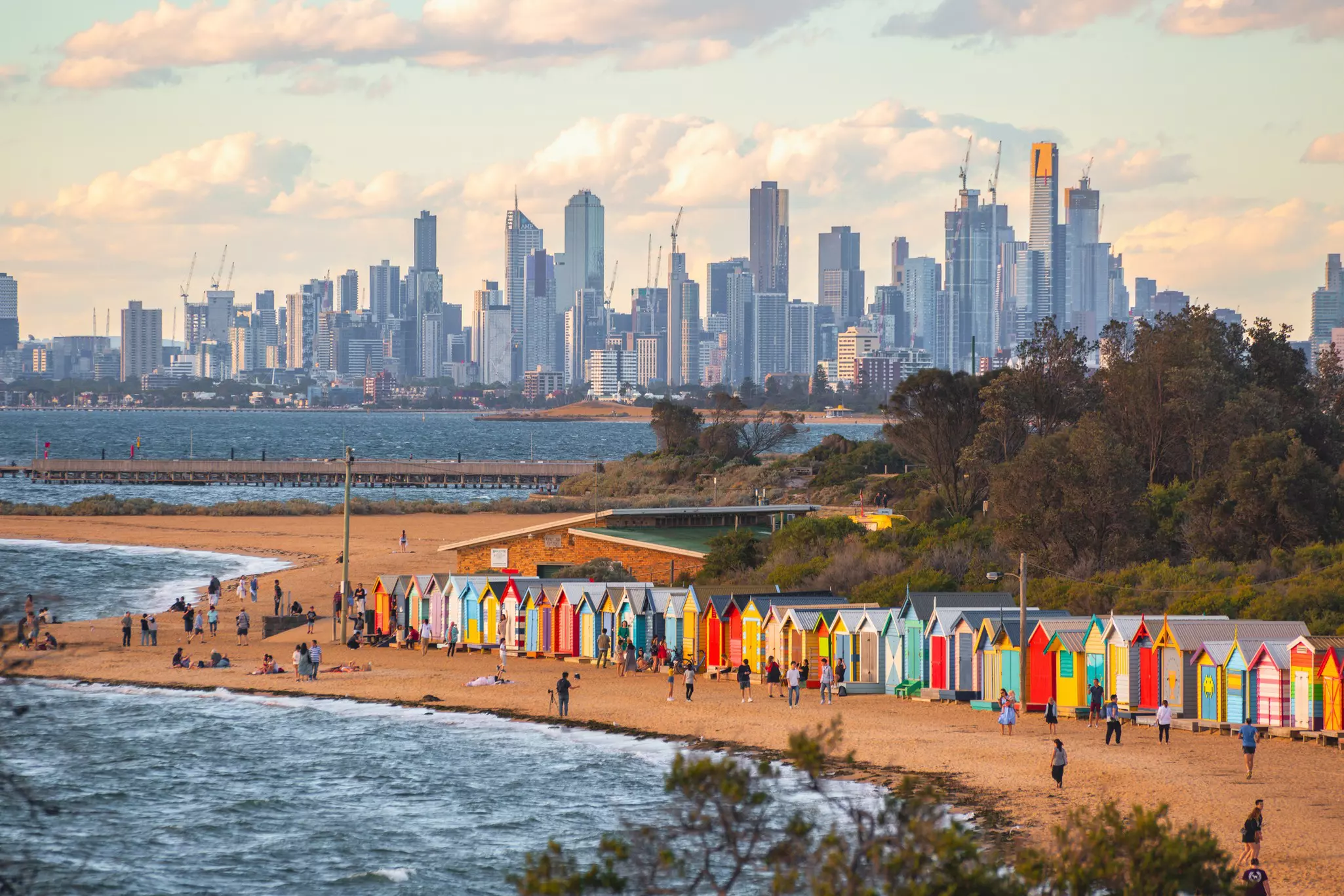 A row of bright colored bathing pavilions at a sandy beach, with the Melbourne skyline on the horizon