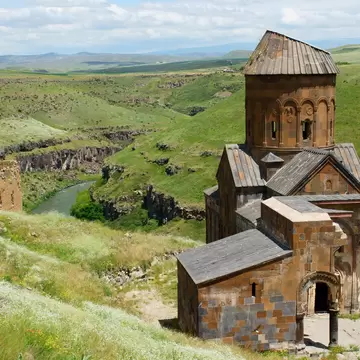 Medieval stone church with a conical-roofed drum on a grassy slope above a green river gorge at Ani, northeastern Turkey.