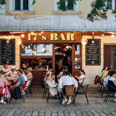 People sitting at outdoor tables in front of a restaurant with the sign for "17's Bar" and blackboards listing the menu items.