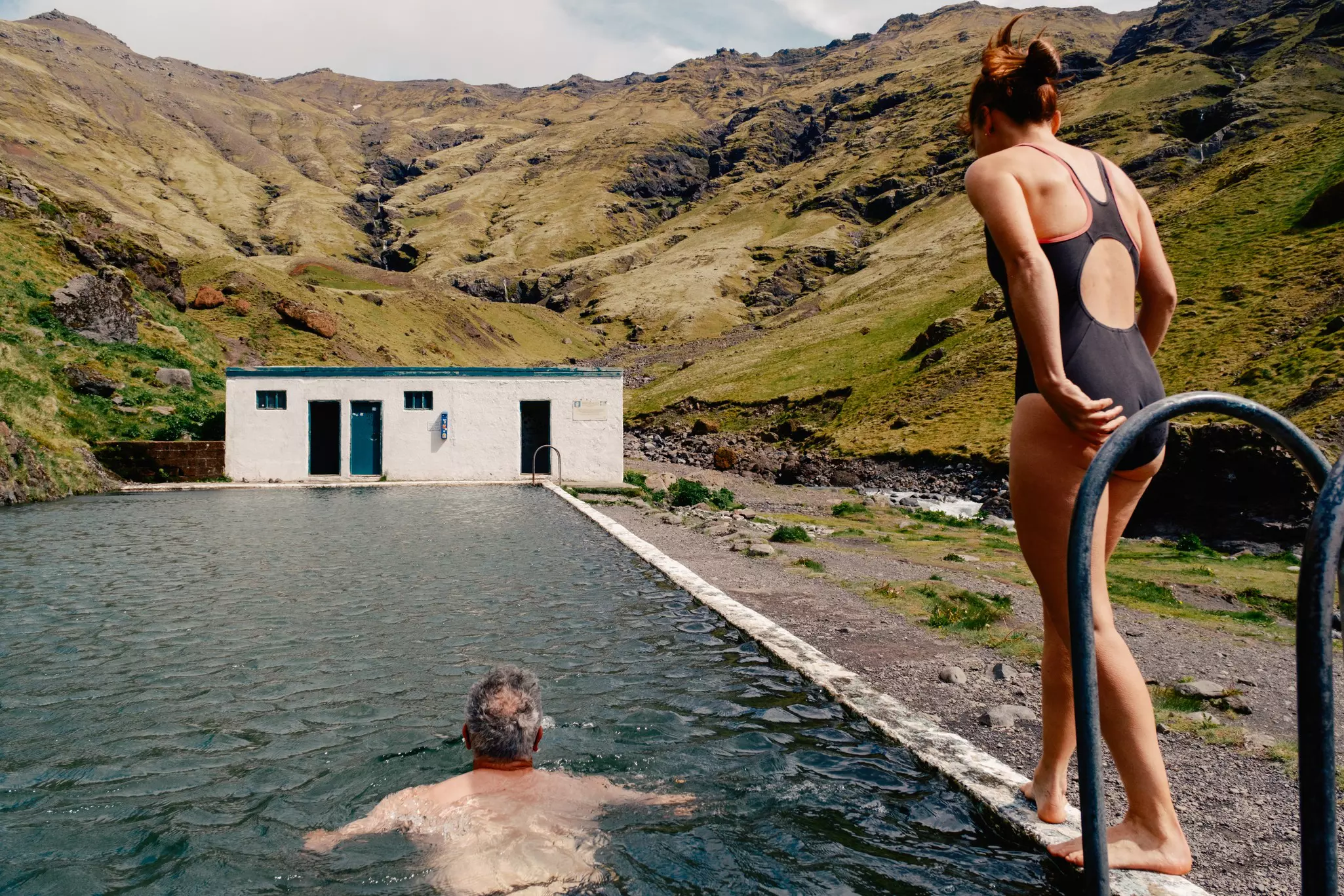 A man swimming as a woman stands on the edge of a pool surrounded by hills in Iceland.