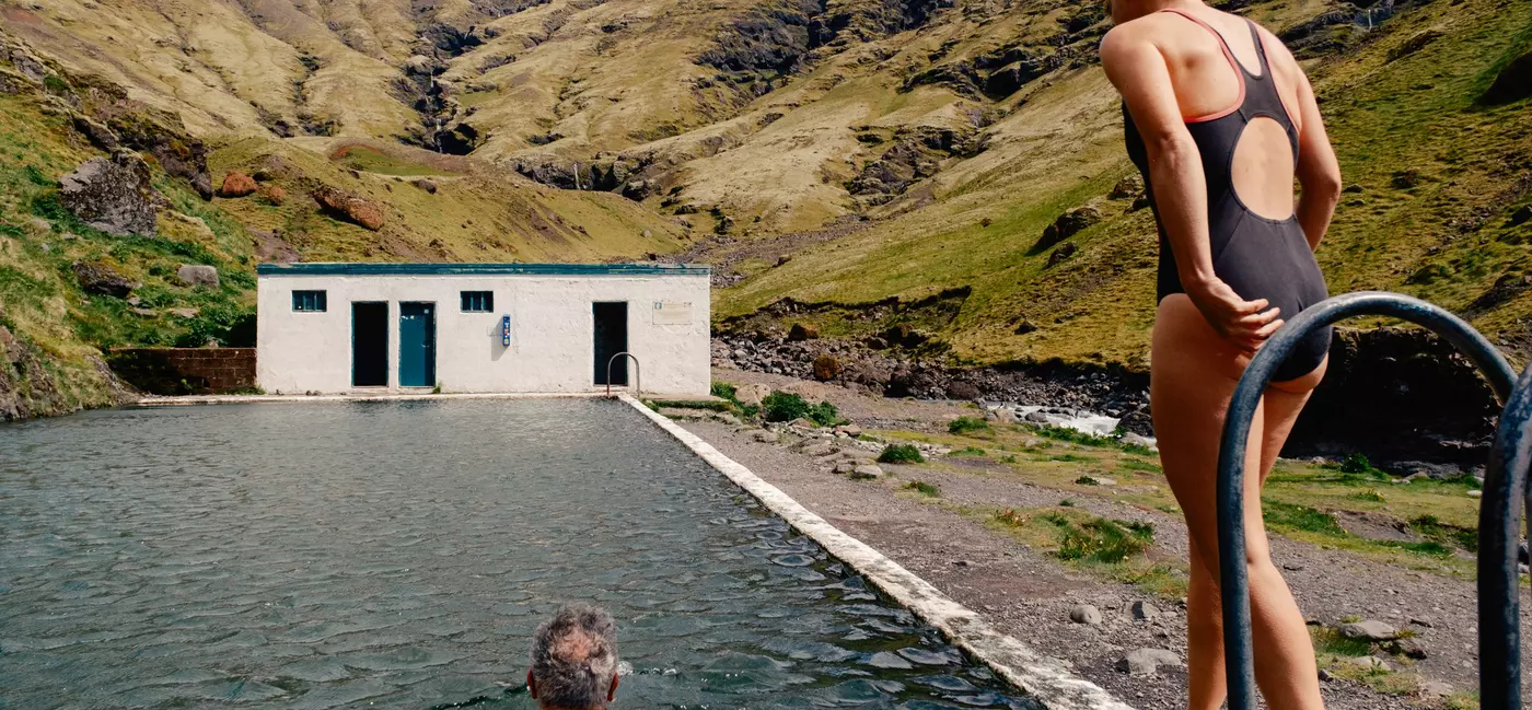 A man swims towards a building in an outdoor pool while a women walks alongside him on his right