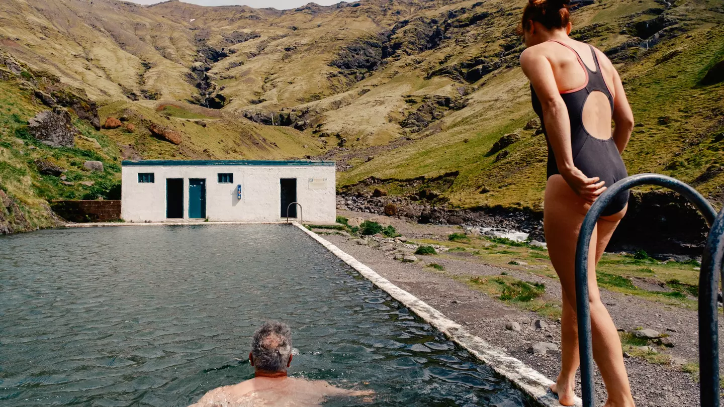 A man swims towards a building in an outdoor pool while a women walks alongside him on his right