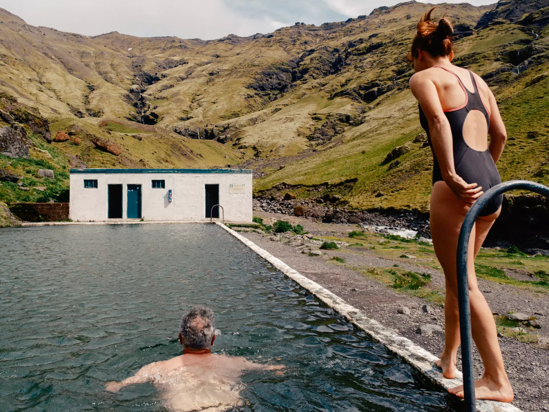 A man swims towards a building in an outdoor pool while a women walks alongside him on his right