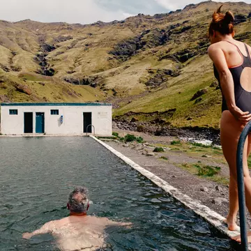 A man swims towards a building in an outdoor pool while a women walks alongside him on his right