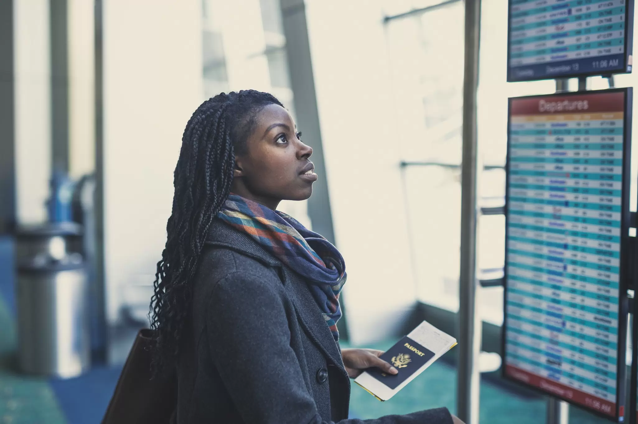 Young woman at an airport holding passport and looking at the departures board