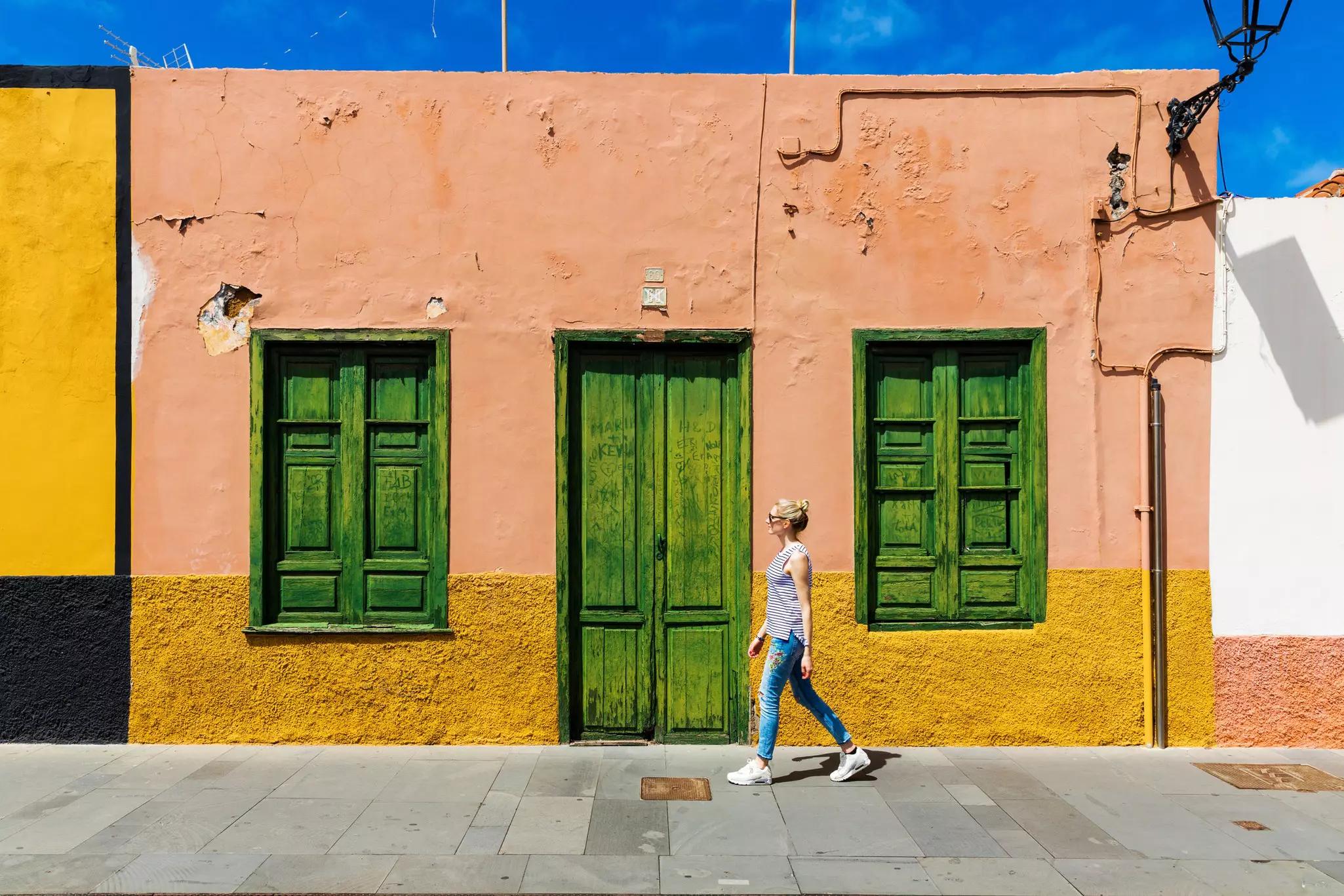 A white woman walks past a single storey pink and yellow house with green shutters in Puerto de la Cruz, Tenerife.