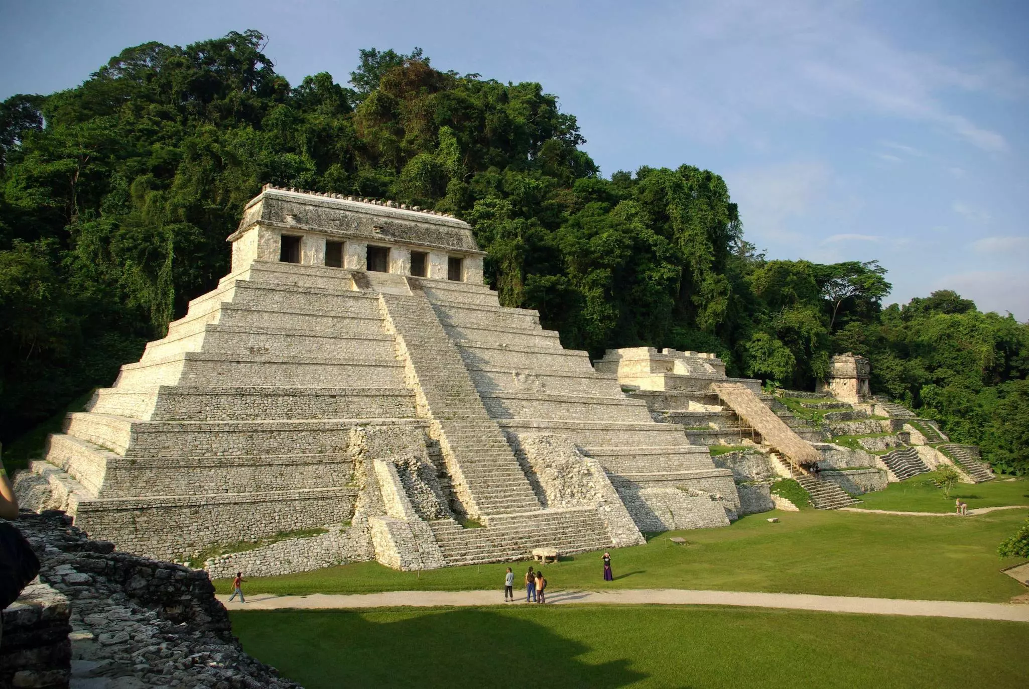 A sand colored stone tiered pyramid with a large front staircase surrounded by jungle and fronting a grass lawn.