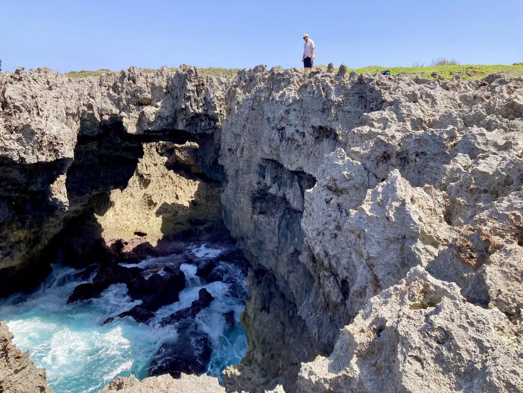 A man stands on a limestone cliff looking down into a cavernous space where waves crash below.