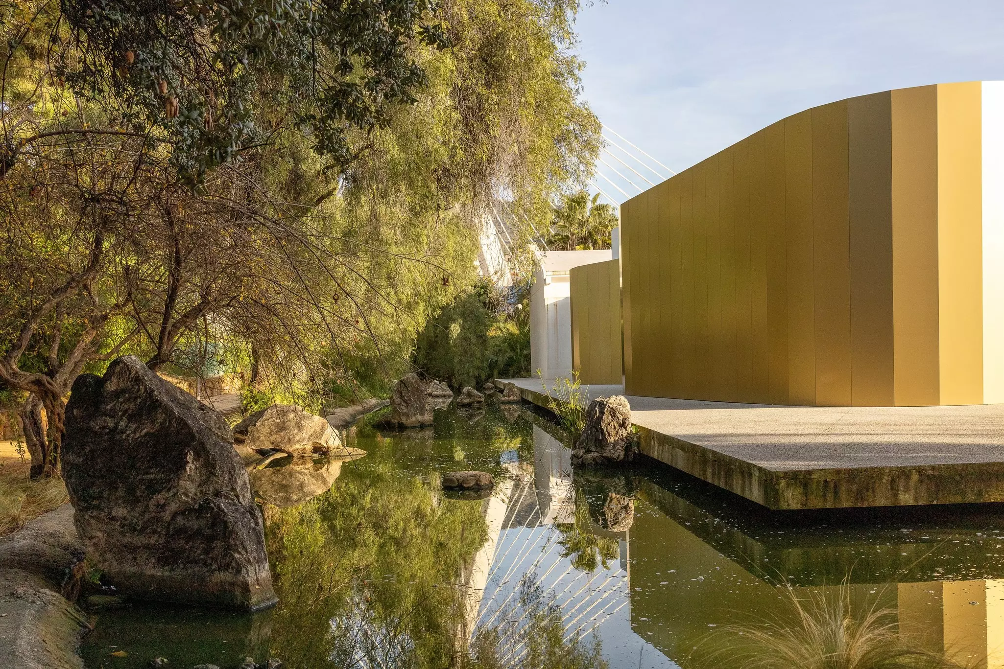 A still water feature with some large rocks surrounds a modern building in a park in Spain.