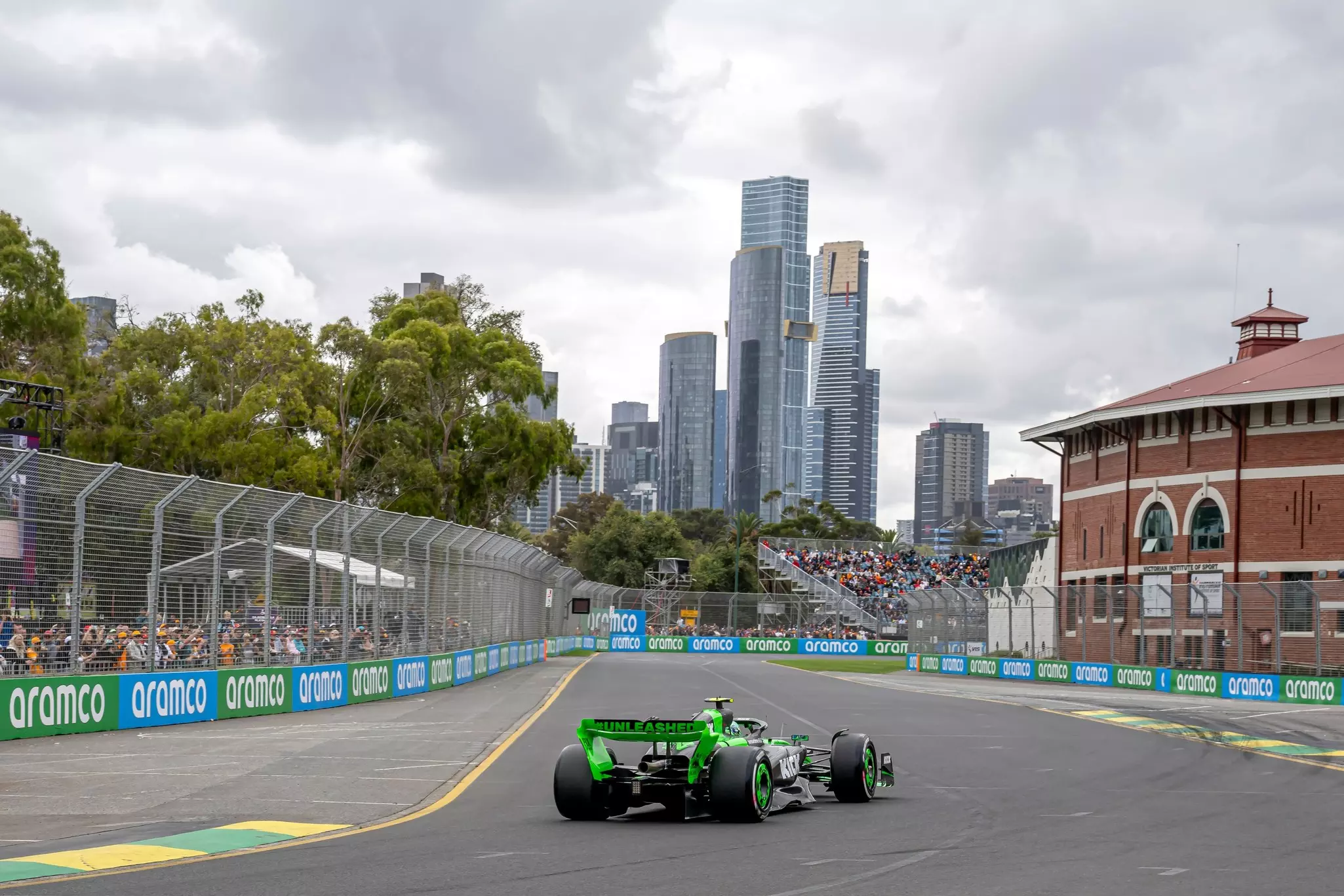 A green racing car on a track within a city with skyscrapers and many spectators in the stands that line the track