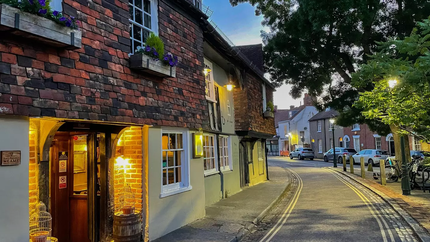 CANTERBURY, KENT, UNITED KINGDOM, SEPTEMBER 14, 2023: Warm glow of a traditional inn shines onto the street on an early summer evening. A charming brick house facade with flower boxes in Canterbury.