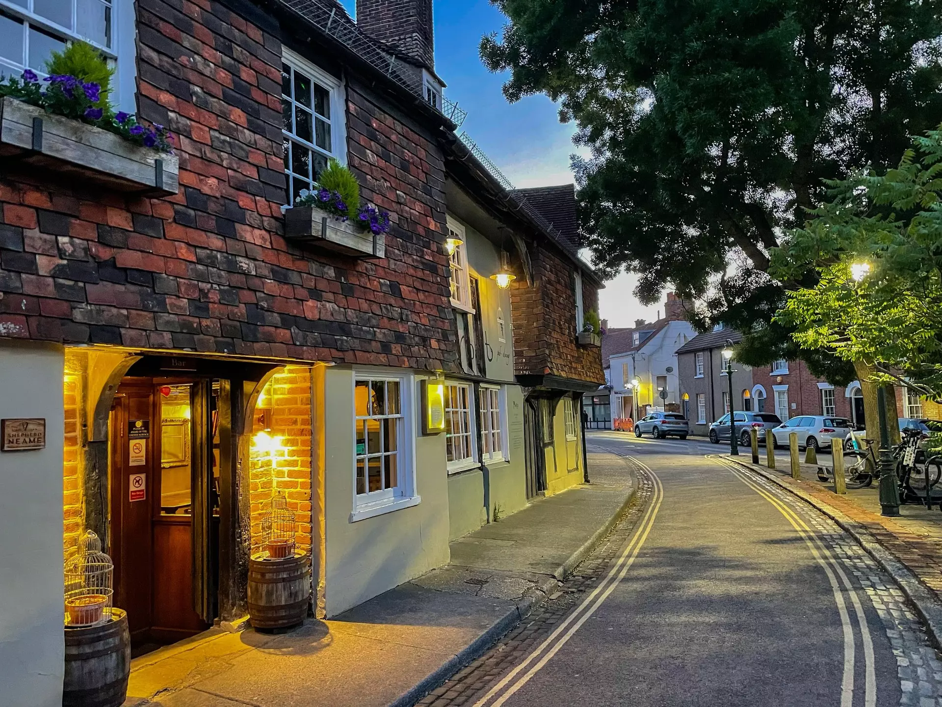 CANTERBURY, KENT, UNITED KINGDOM, SEPTEMBER 14, 2023: Warm glow of a traditional inn shines onto the street on an early summer evening. A charming brick house facade with flower boxes in Canterbury.