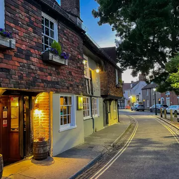 CANTERBURY, KENT, UNITED KINGDOM, SEPTEMBER 14, 2023: Warm glow of a traditional inn shines onto the street on an early summer evening. A charming brick house facade with flower boxes in Canterbury.