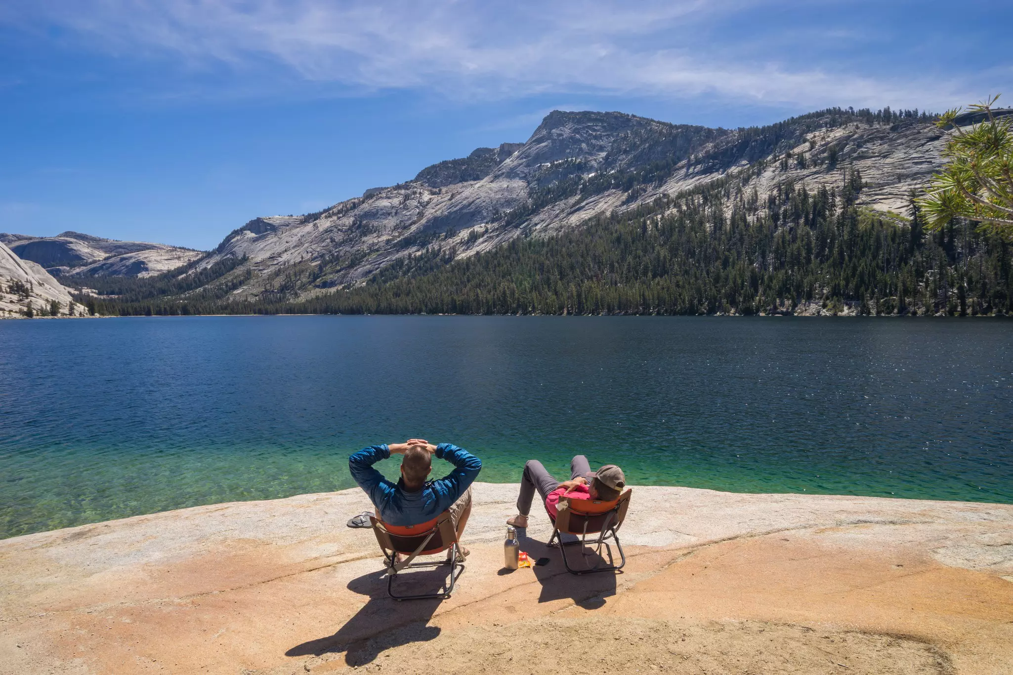 Two people sitting in camp chairs at the edge of a lake facing mountains.