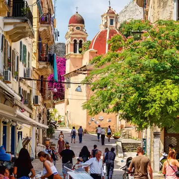 Sloping stone lane with pedestrians and cafe tables, tall shuttered buildings and a pink church with red domes, Corfu Town.