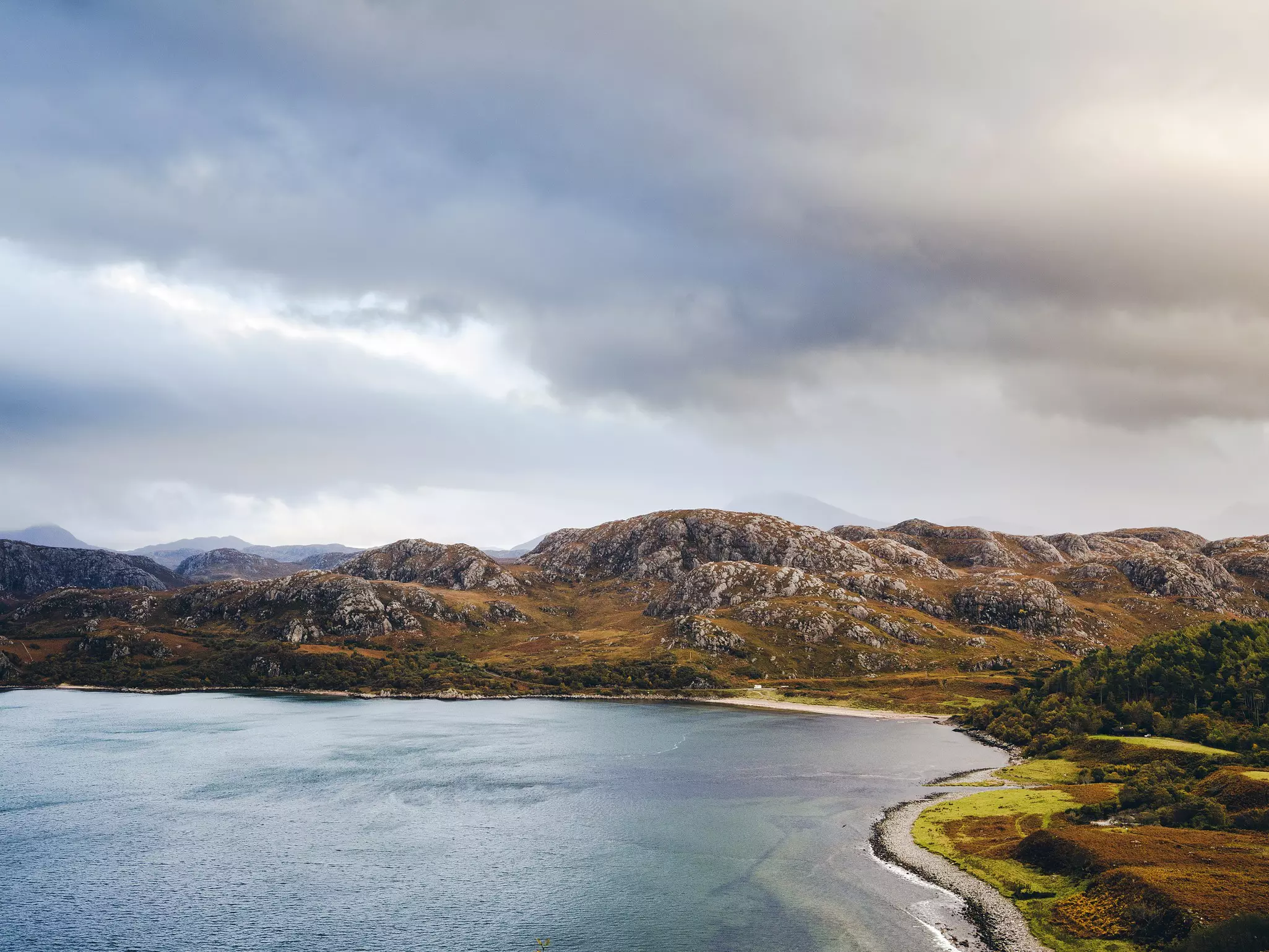 One of the westernmost stretches of the NC500 passes by a small beach at Gruinard Bay. An image of the bay with trees on the right and mountains in the background, under a cloudy sky.