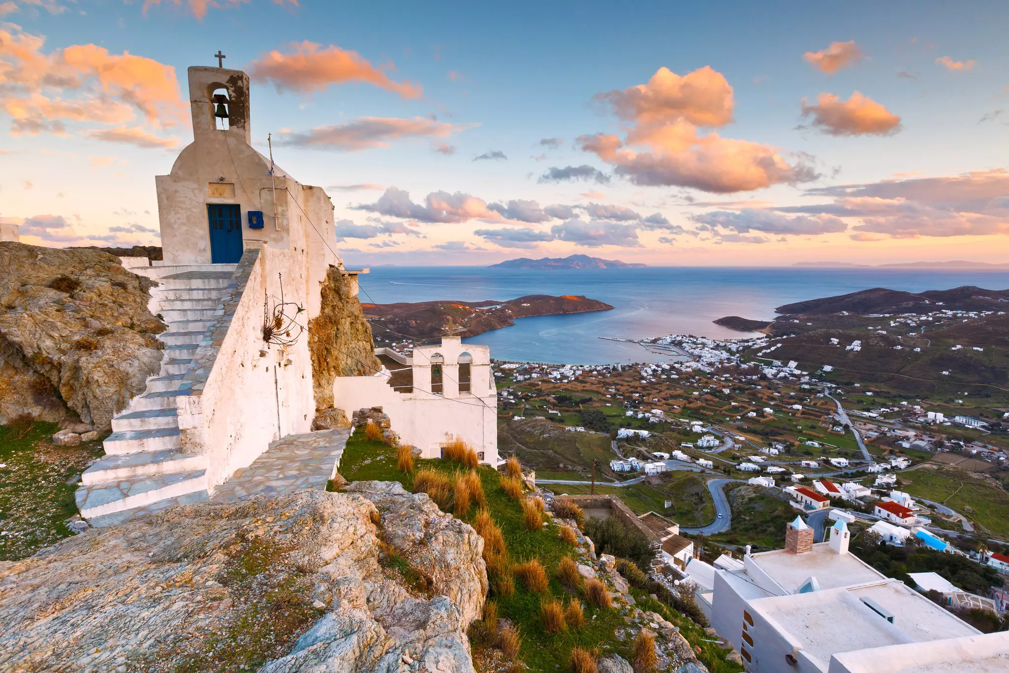 Steps lead to a narrow white building on top of a steep hill; below, scattered white buildings radiate out from a harbor in the Cyclades, Greece.