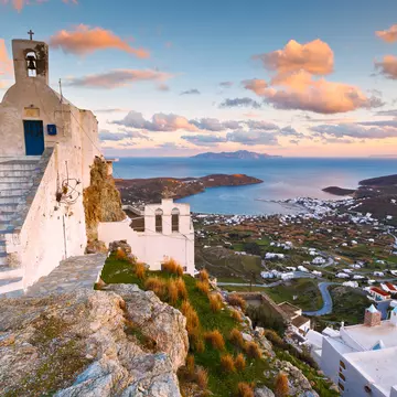 Steps lead to a narrow white building on top of a steep hill; below, scattered white buildings radiate out from a harbor in the Cyclades, Greece.