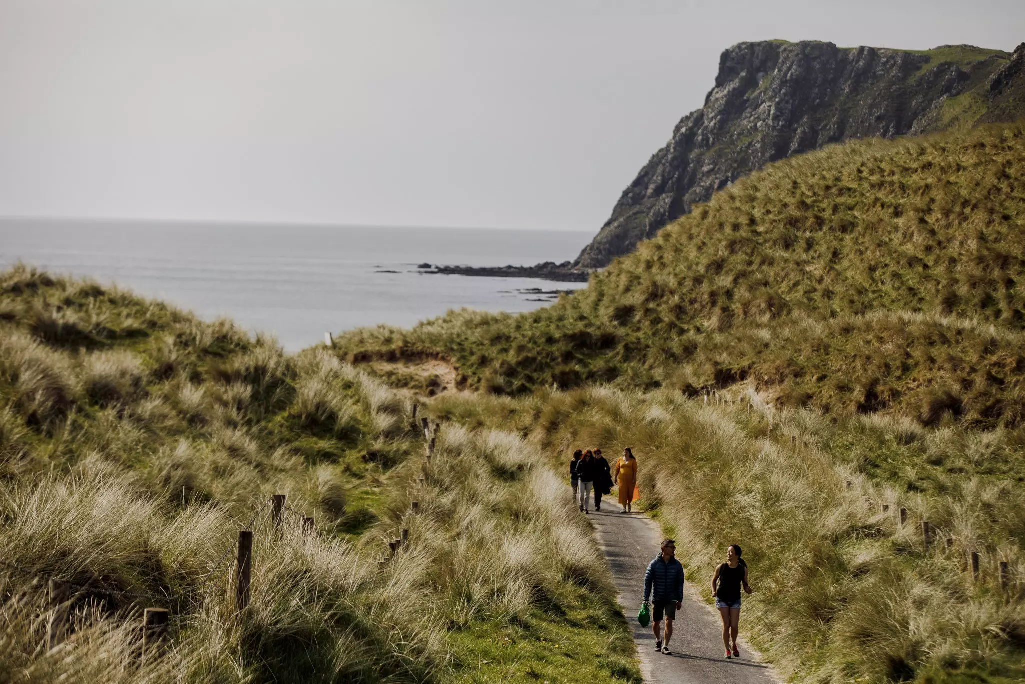 People walk on a boardwalk through grassy hills toward the ocean.