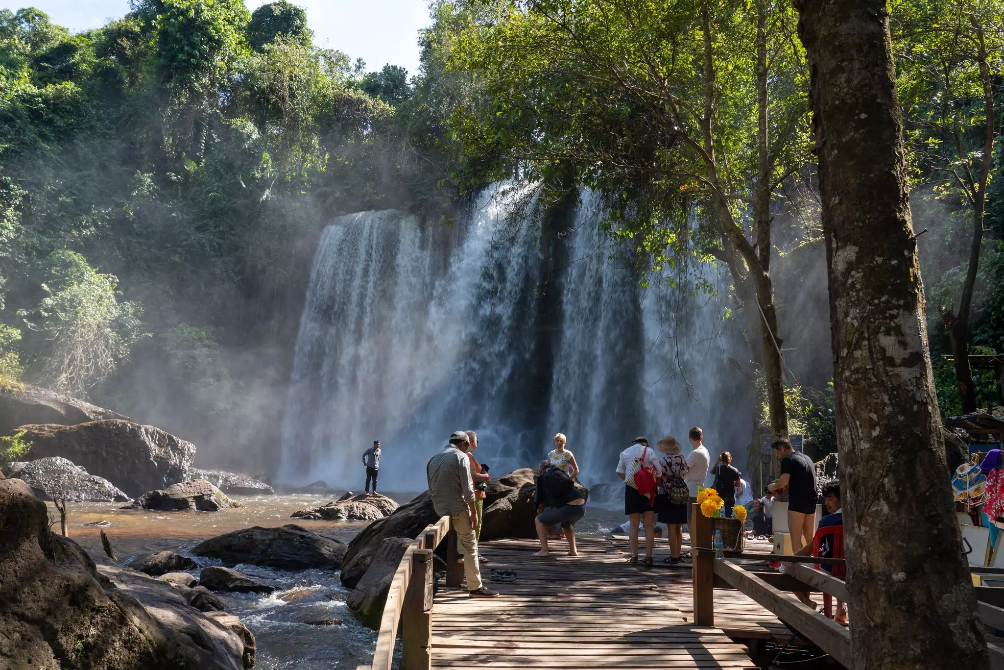 Visitors stand on a wooden platform looking toward a waterfall at Phnom Kulen in Cambodia.
