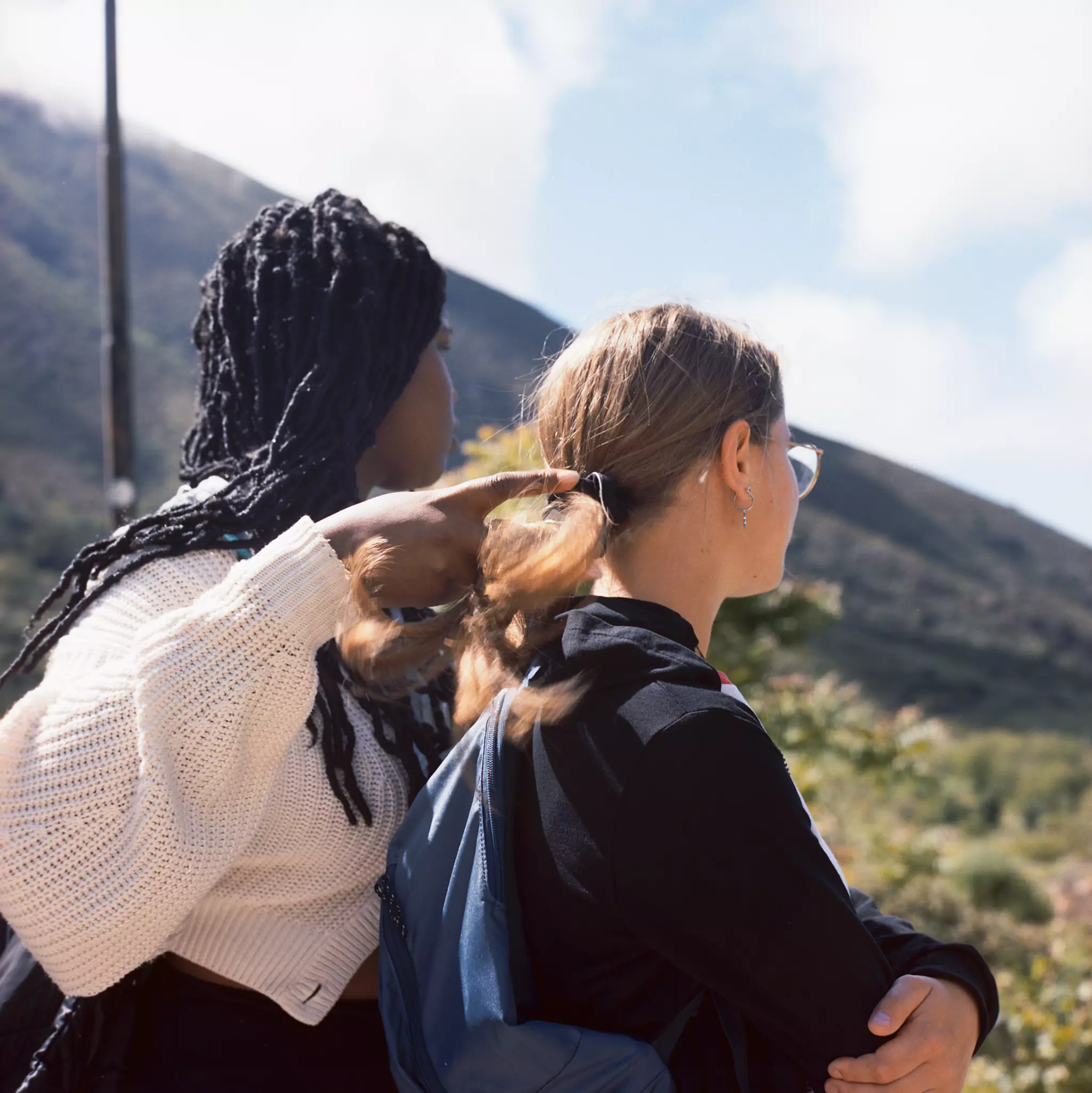 Two young woman sitting next to one another on a sloped field are seen from behind, in partial view.