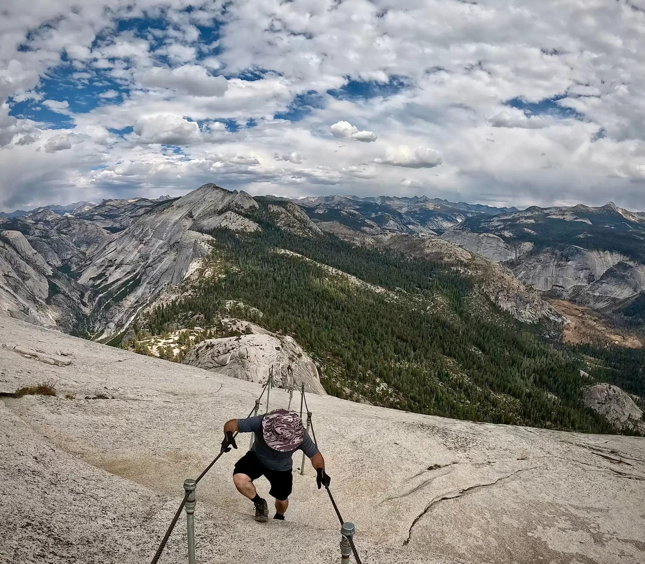 A hiker pulls himself up the very steep incline of a granite dome using fixed cables. The mountains disappear into the distance: the cable route up Half Dome in Yosemite, California.