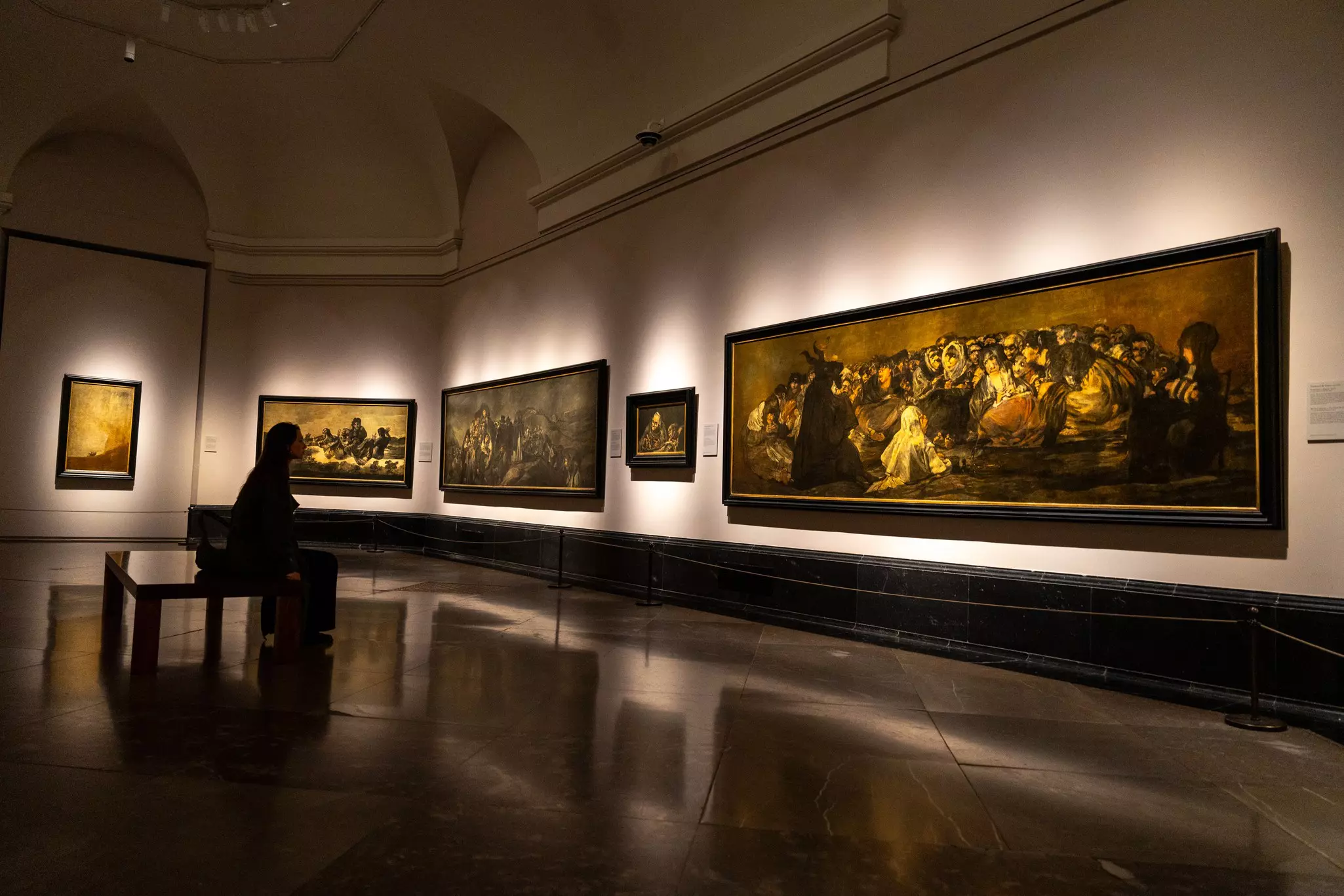 A person sits on a bench in a darkened gallery looking at large paintings on a wall.