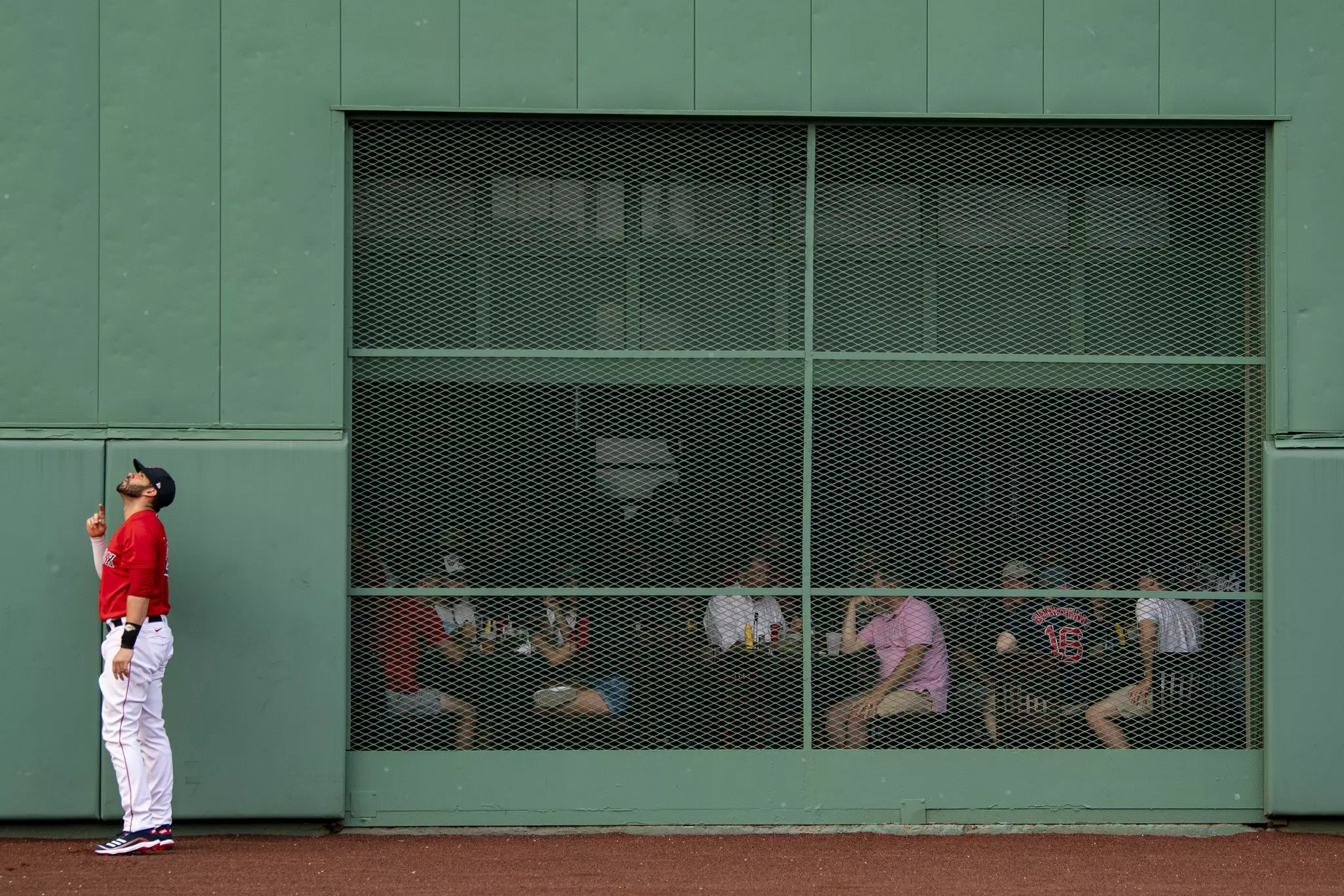 A baseball player in a red uniform looks up on the field in front of a green wall. People sitting at a bar are seen behind a grated window, looking out at the field.