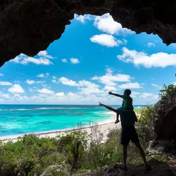 Seen from inside a rocky cave mouth, a silhouetted father and child point at birds over a white beach and turquoise sea.