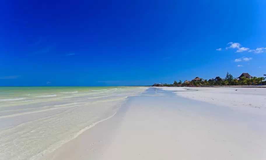 Wide empty white-sand beach with shallow turquoise water and distant thatched buildings