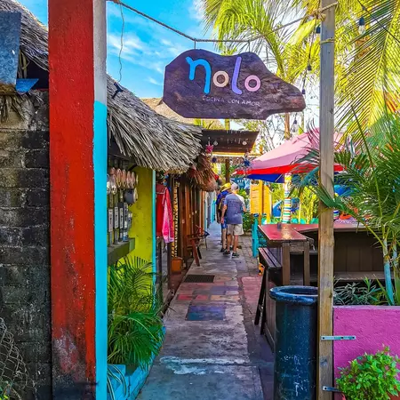 A narrow sidewalk with a restaurant sign above and seating to the right. People walk in the distance on a sunny day.